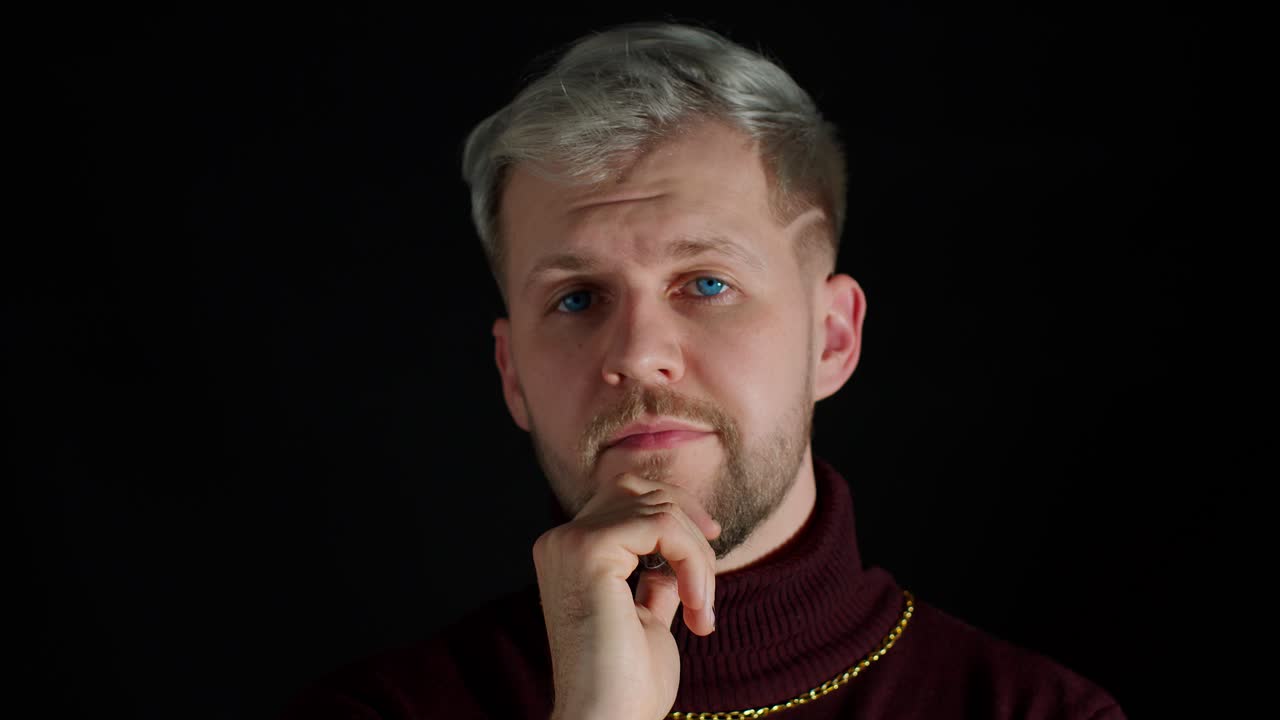 Cheerful thinking young man holding hand on chin looking at camera and smiling on black background