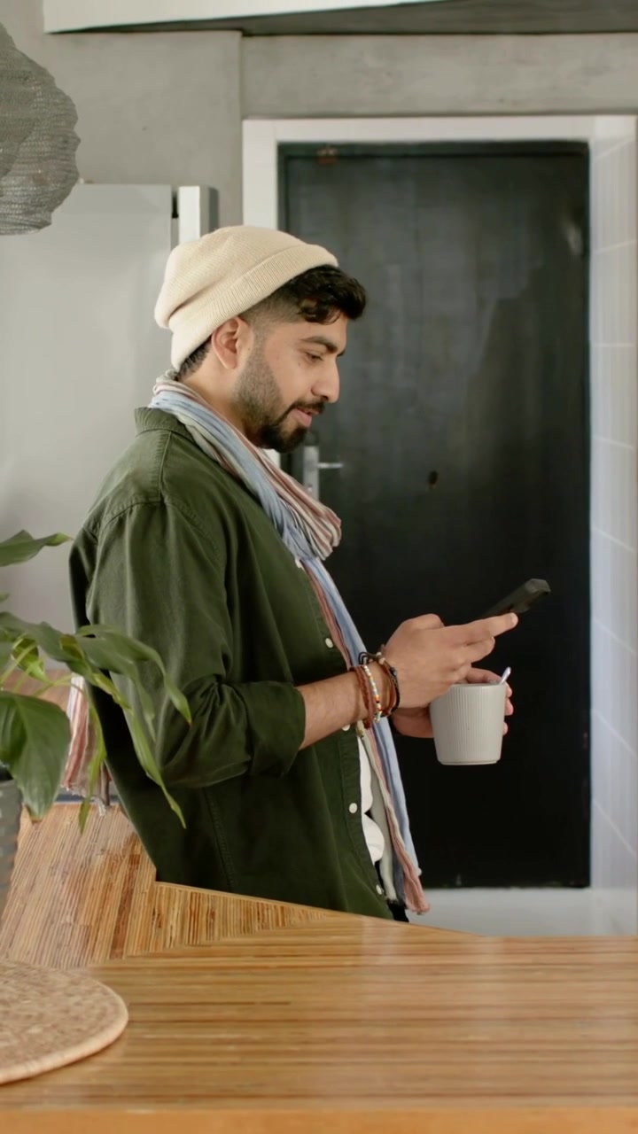 Vertical video: Reaching for phone, beanie-clad man picking it up in kitchen holding mug reading