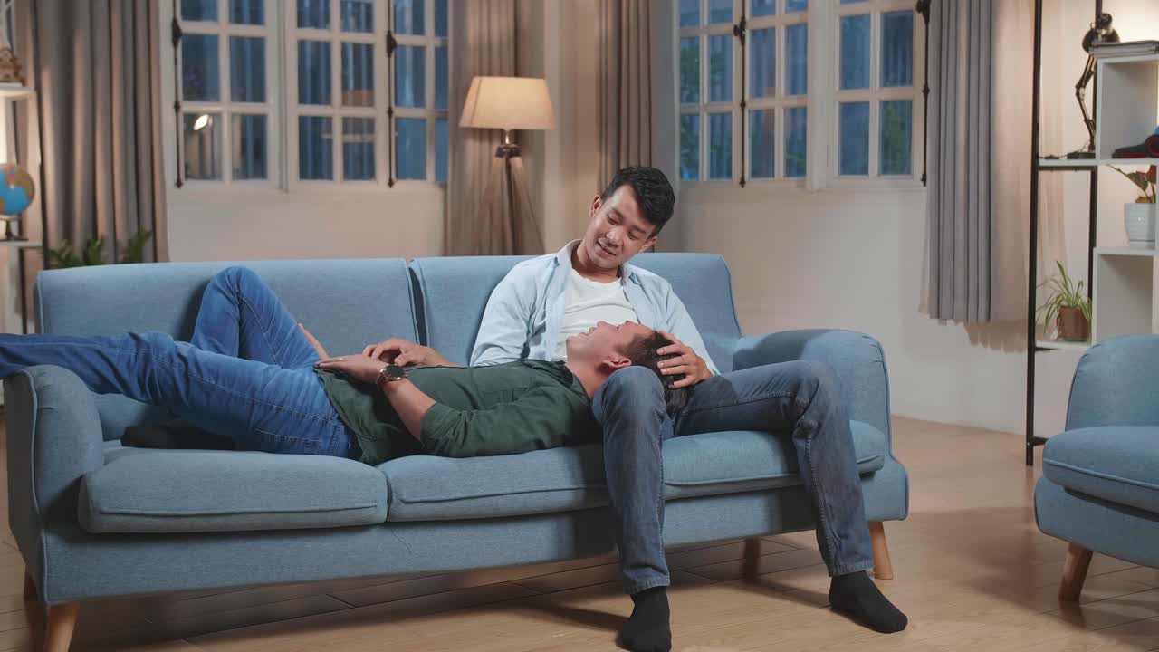 Young Asian Gay Couple Spend Time At Home. Two Happy Man In Love Lie Down On Sofa And Cuddle In Casual Clothes. Cute Lgbt Relationship Content