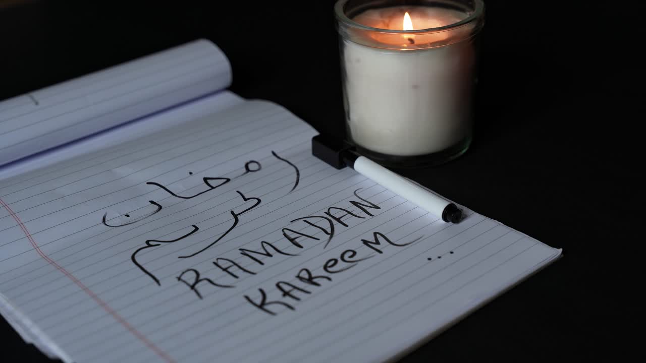 Primo piano di una mano che scrive Ramadan Kareem in arabo e inglese su un quaderno a righe, accanto a una candela accesa, scena interna con poca luce per gli auguri delle festività islamiche.