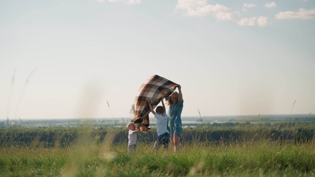 A woman in a flowing blue dress joyfully holds a plaid scarf that dances in the breeze on a lush grassy hilltop. Two children in white shirts eagerly reach out to touch the scarf
