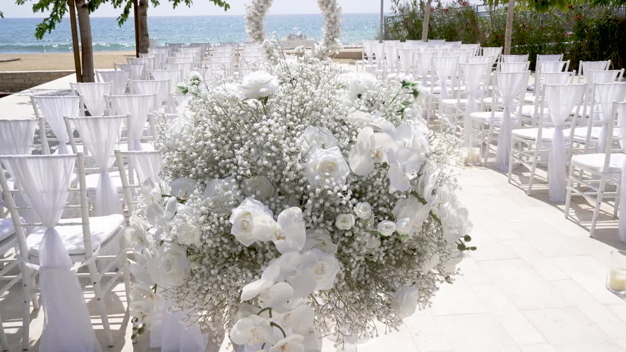 Un pittoresco allestimento per un matrimonio sulla spiaggia include file di sedie bianche e sontuose decorazioni floreali. Il cielo limpido e l’oceano sullo sfondo creano un’atmosfera romantica.