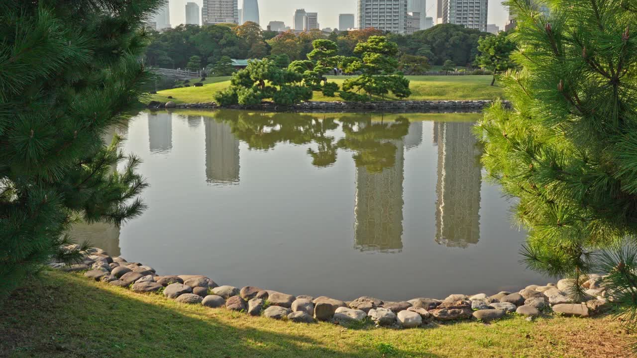 A tranquil pond in Hamarikyu Gardens reflects modern city buildings under a clear sky, framed by green trees and a rock-lined shore.