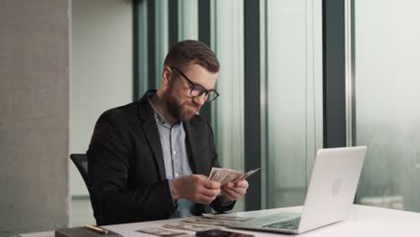 Un-Joven-Feliz-Con-Ropa-De-Negocios-Cuenta-Billetes-De-Dinero-Mientras-Está-Sentado-Frente-A-Una-Laptop