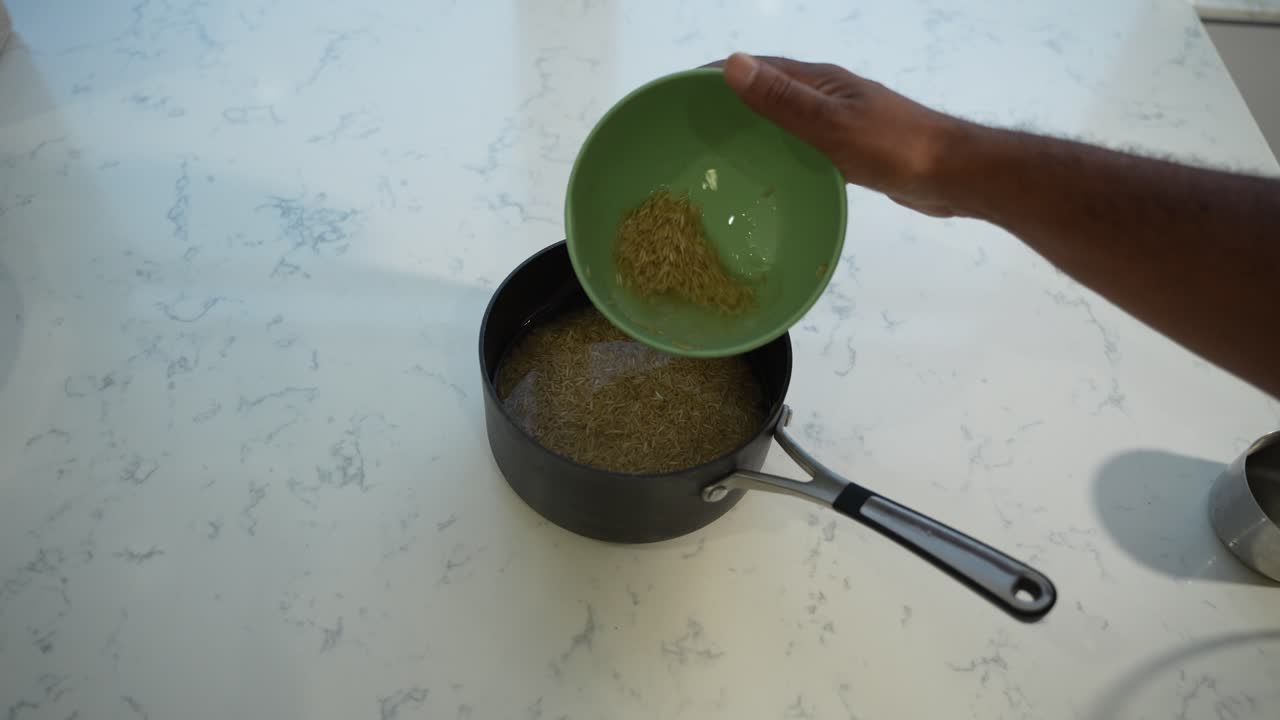 A hand pours dry rice into a black pot on a marble countertop, preparing a meal in a kitchen setting