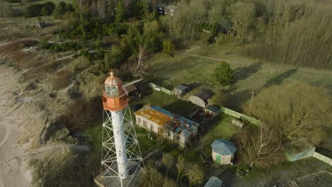 Aerial-establishing-view-of-white-colored-Pape-lighthouse,-Baltic-sea-coastline,-Latvia,-white-sand-beach,-large-waves-crashing,-sunny-day-with-clouds,-wide-drone-shot-moving-backward,-tilt-up