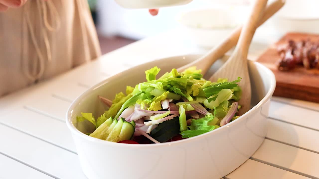 A person prepares a vibrant vegetable salad using tongs and wooden spoons in a well-lit kitchen setting
