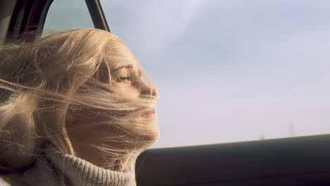 woman looks out open car window as blond hair swirls across face, close-up