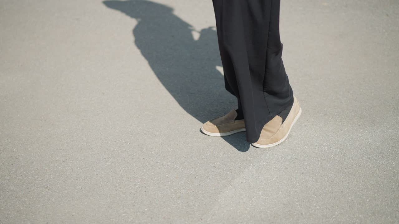 Individual stands quietly on sunlit pavement wearing black wide pants and brown shoes, strong shadow stretches across ground, symbolizing calm movement, curiosity, and thoughtful stillness