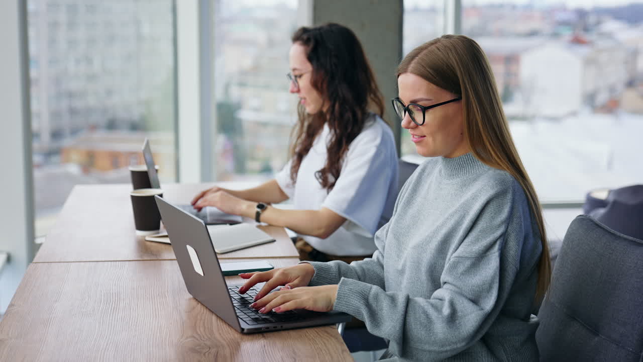 Two positive ladies work at laptops in the office. Women wearing computer glasses type and talk.