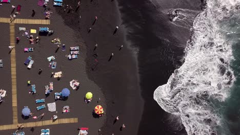 Aerial-top-down-of-beach-summer-vibes-scene-with-people-sunbathing-and-ocean-waves-in-black-sand-tropical-beach