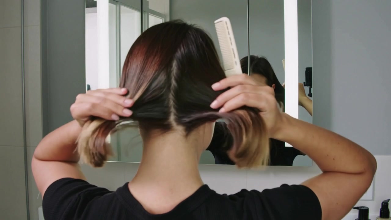 Mujer con top negro mirándose al espejo, peinándose el cabello en el lavabo del baño con un peine blanco, con un botiquín.
