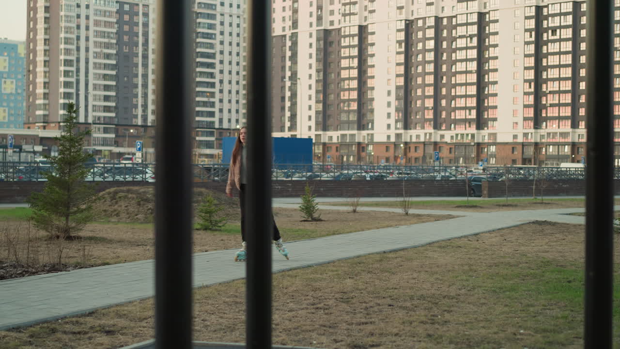 A young girl wearing a peach jacket and black trousers is rollerblading along a paved path in an urban park. The scene features tall buildings, parked cars, and an iron pole in the background