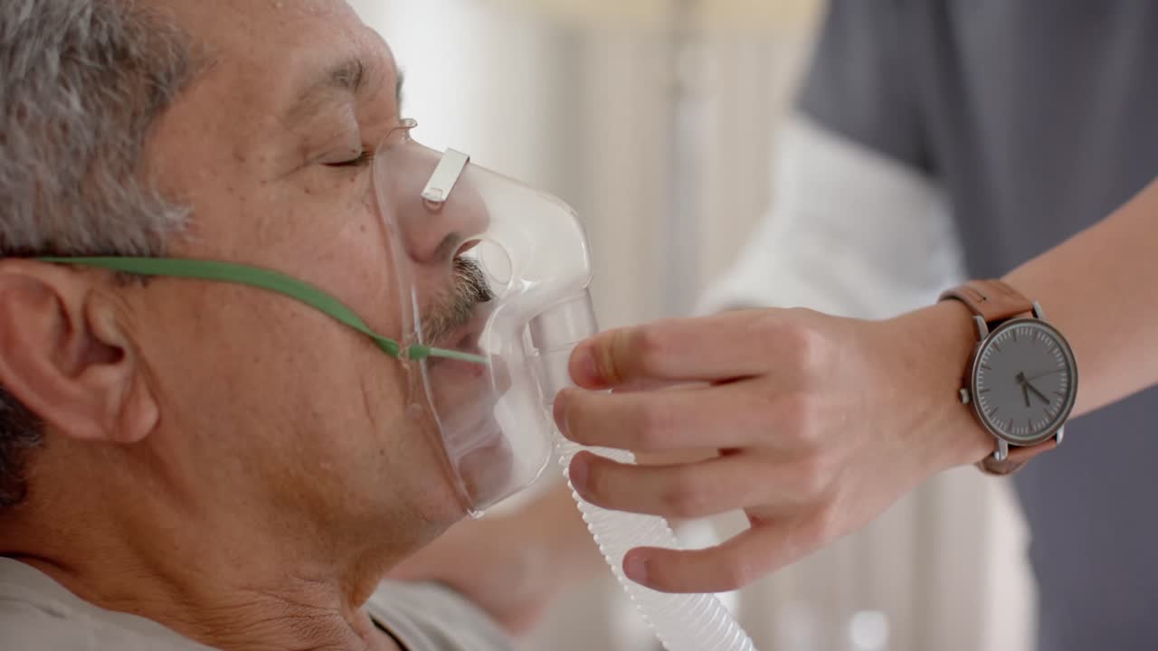 médico varón diverso ayudando a un paciente varón anciano usando una máscara de oxígeno en casa, cámara lenta