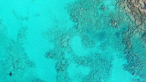 Drone-view-in-Greece-top-view-ocean-clear-blue-water-with-rocks-on-the-bottom-a-small-white-boat-on-a-sunny-day