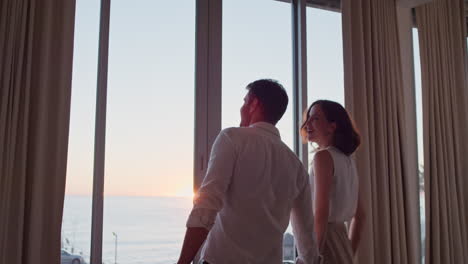 happy travel couple arriving in hotel room on honeymoon vacation looking out window enjoying beautiful view of ocean at sunset embracing sharing romantic holiday