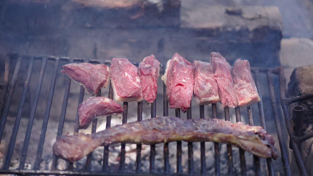 Slow camera slide over Argentine asado grill with skirt steak (entraña) and Top Sirloin Cap ( picaña ) cuts grilling above wood embers, smoke rising and meat sizzling in authentic outdoor barbecue