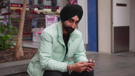 Young-Indian-Man-Using-Phone-Looks-To-The-Camera-And-Smiles