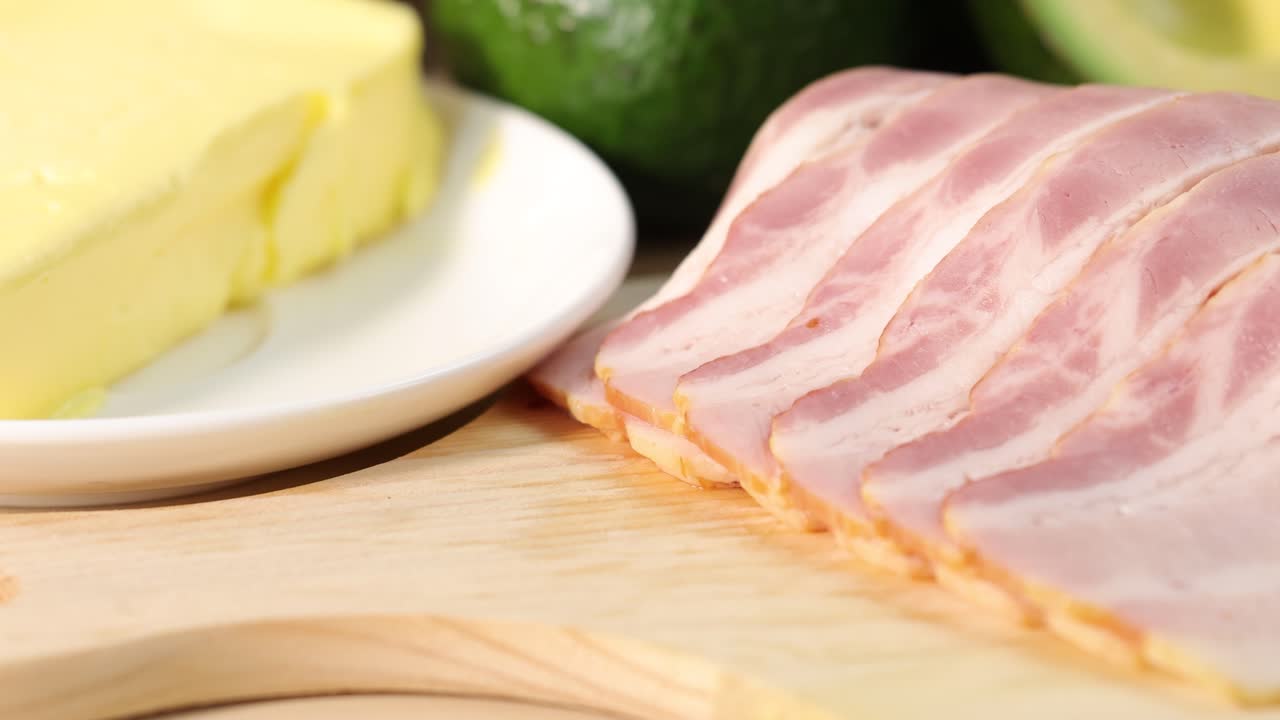 A smooth pan across nuts, seeds, butter, and bacon slices on a wooden surface under bright, natural lighting, emphasizing healthy fat sources and food textures