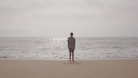 boy standing on a beach, watching the waves