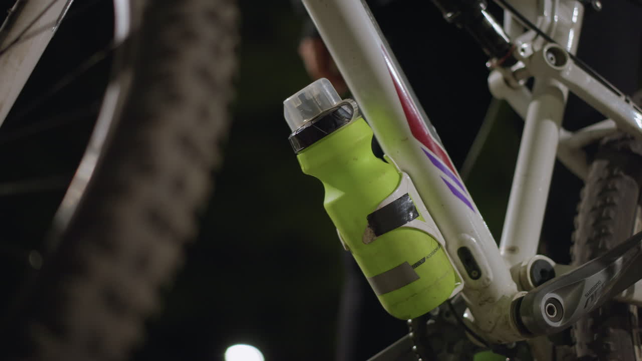 Closeup Of Rider Hand Refilling Neon Green Bottle Attached To Mountain Bike Frame, Soft Park Lights In Background, Textured Tire And Suspension Visible, Purposeful Motion And Hydration Focus