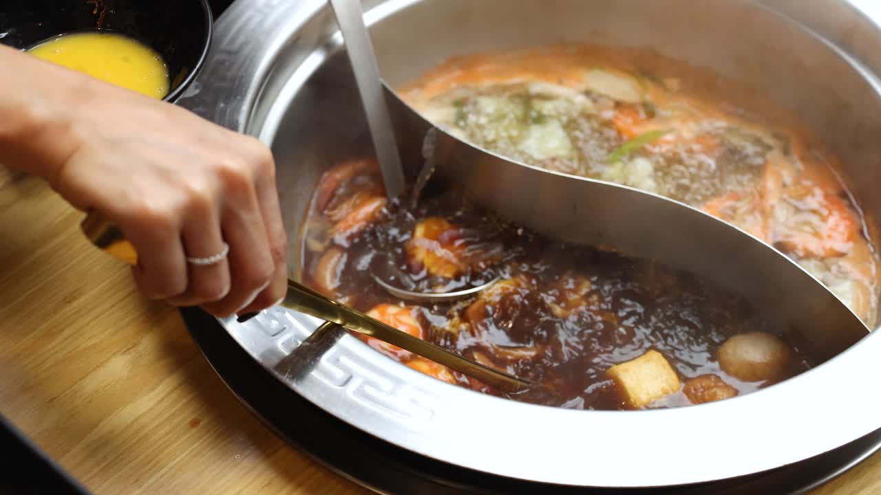 Hand cooks beef slice in steaming dual broth hotpot, tabletop view, warm lighting, minimal movement