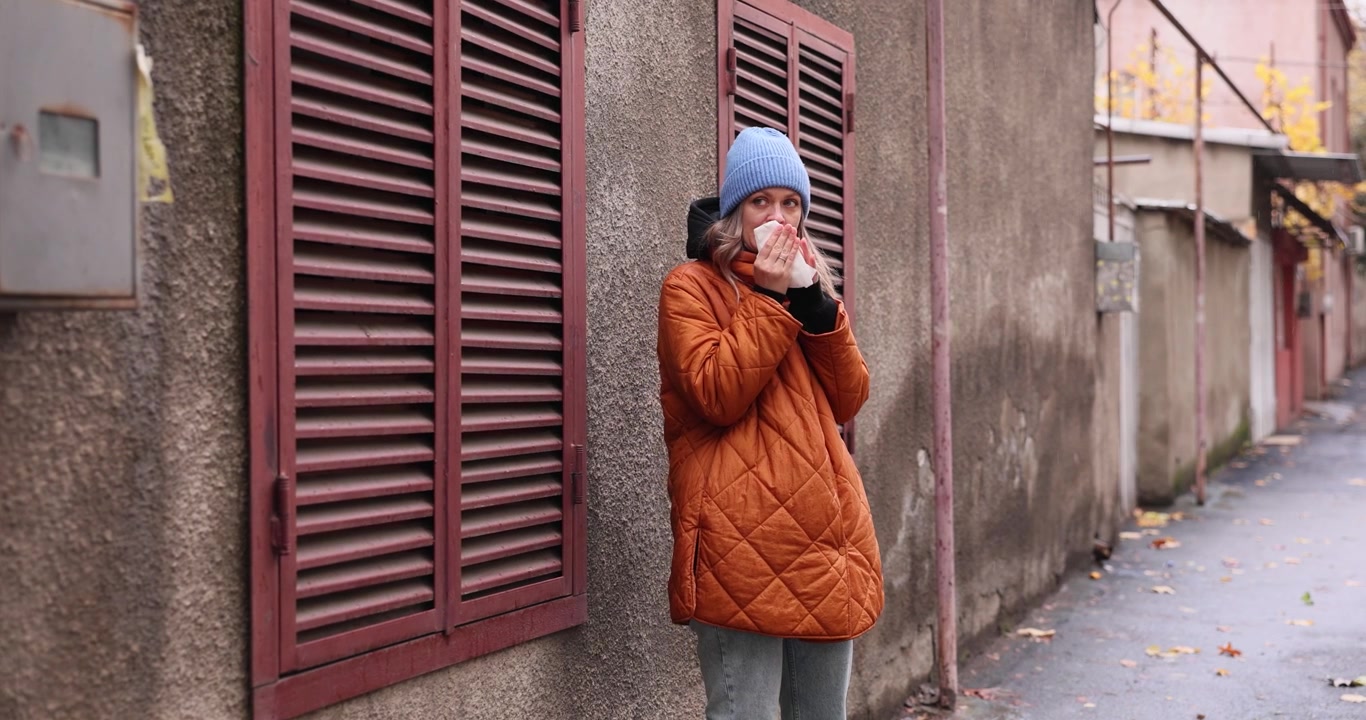 Donna con giacca e cappello si soffia il naso in piedi vicino a un vecchio edificio