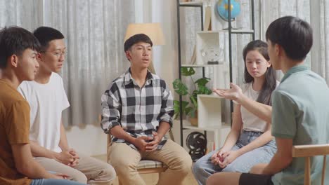 아시안 십대 그룹은 의자에 앉아서 집에서 토론하는 원을 형성합니다. 회색 셔츠를 입은 소년이 친구들에게 동기를 부여하고, 코칭하고, 프로젝트를 계획하고 있습니다.