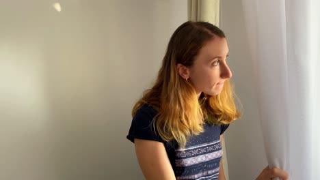 mujer joven camina a la ventana y sostiene la cortina buscando curiosidad mientras observa afuera