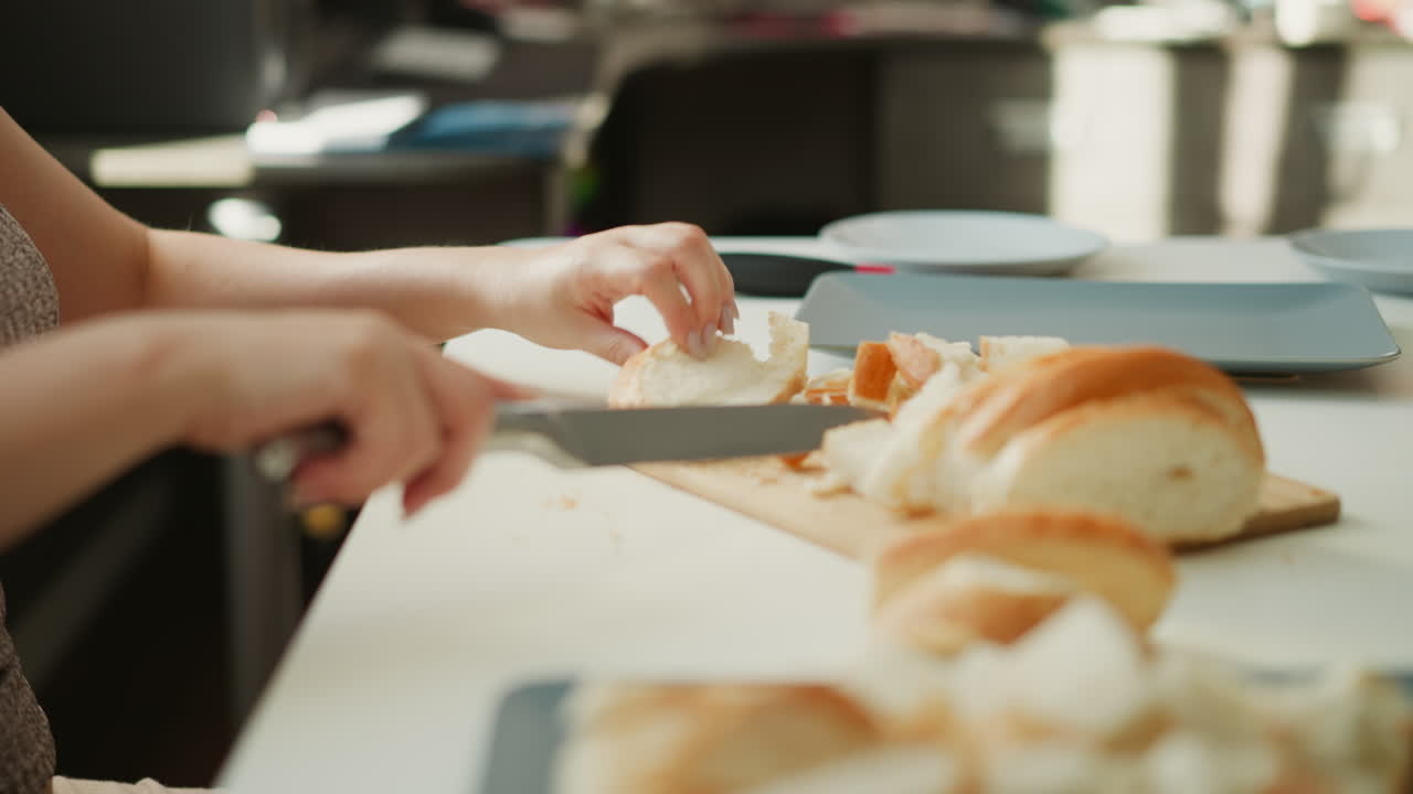 Soft window light illuminates side view of young chef hands slicing loaf into small bite sized pieces on white table with crumbs scattered, capturing focused calm culinary preparation moment
