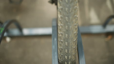 close-up front view of bicycle tire secured in metal bike rack, highlighting frame and spoke details against coal tar pavement with soft shadows cast across concrete