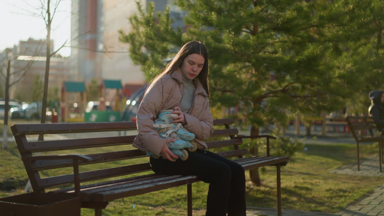A young person in a peach jacket and black trousers sits on a park bench in the warm sunlight, holding a pair of blue rollerblades. The background features greenery, people, and playground equipment