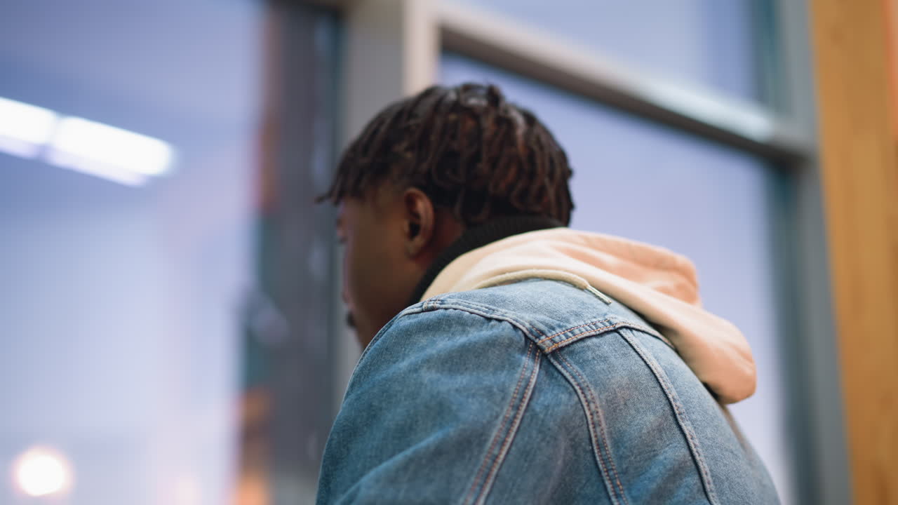 Young man in casual denim jacket and layered hoodie drinks hot beverage from paper cup while seated indoors near window, focused and calm, with soft light illuminating
