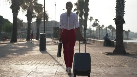 wide shot back view of gorgeous slim businesswoman walking with purse and travel bag in slow motion outdoors. confident caucasian lady strolling on mediterranean embankment. business trip concept.