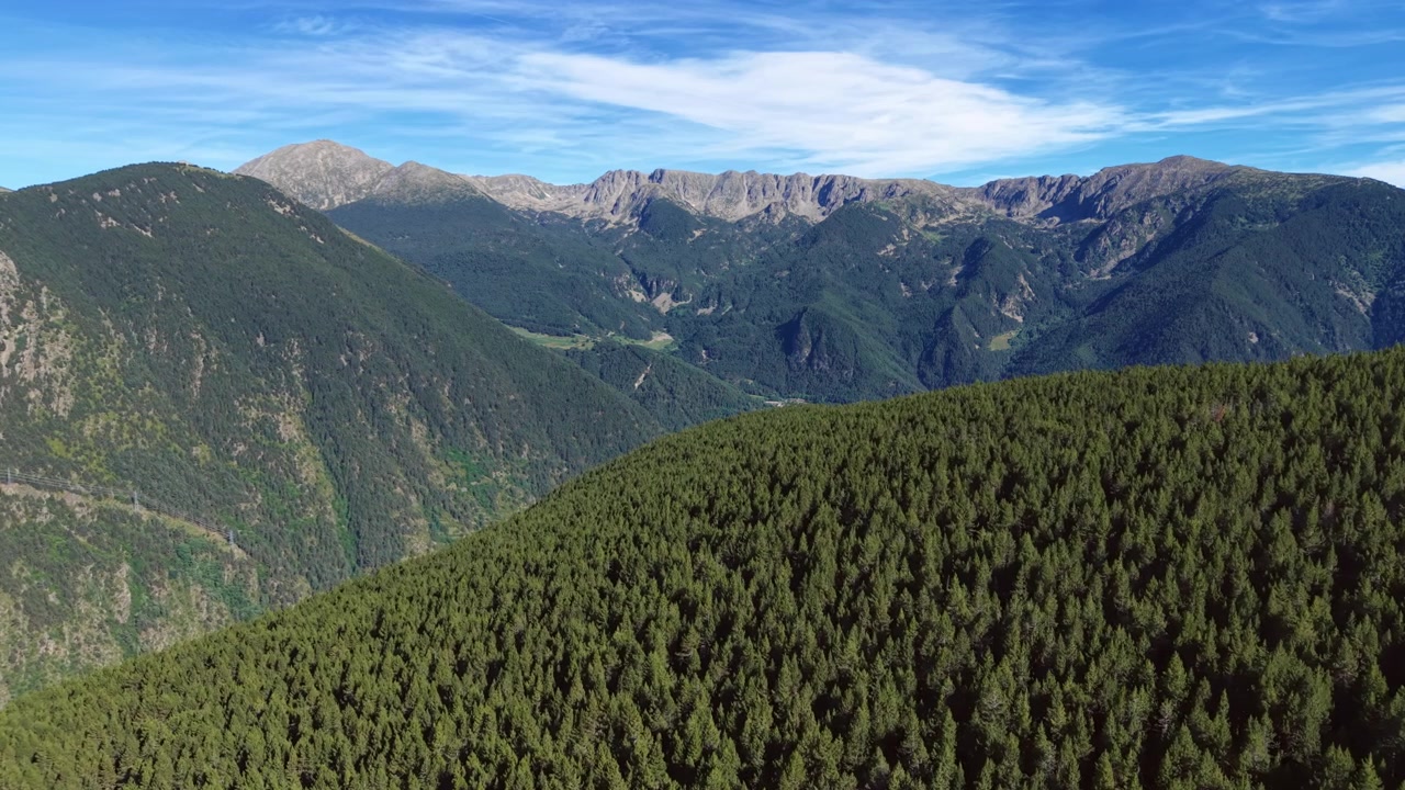 Pemandangan panorama pegunungan Pyrenees Andorra dengan hutan pinus lebat menutupi perbukitan landai, puncak berbatu, dan lembah hijau di bawah langit biru dengan awan tipis