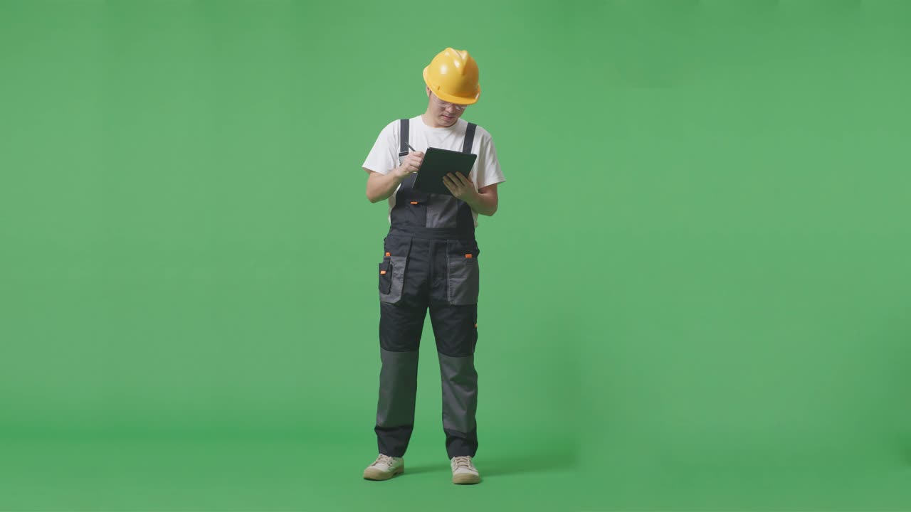 полное тело азиатского мужчины рабочий в защитных очках и защитном шлеме делает заметки на планшете, стоя в зеленом экране фоновой студии