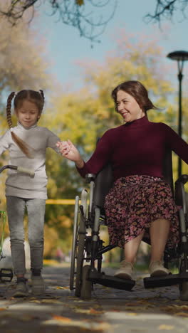 young aunt and preschooler niece spend time walking together with smile. gentle girl supports woman in wheelchair holding hand lovingly. recreation in park