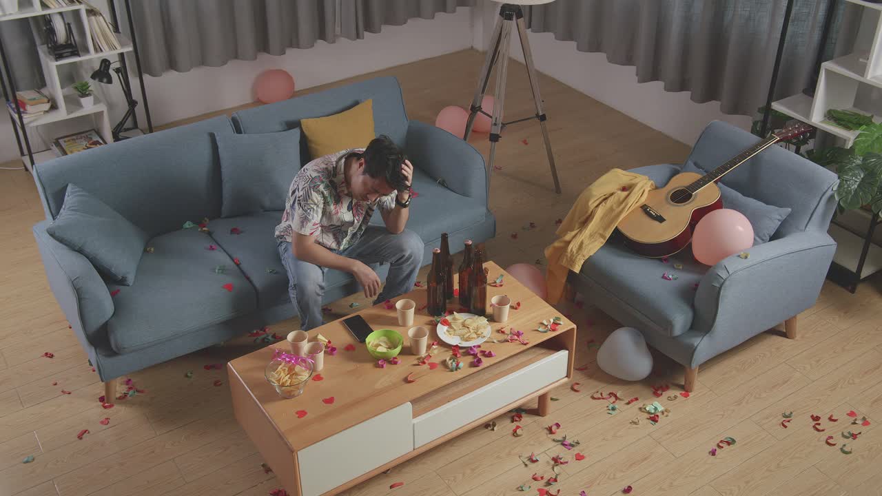 vista superior de un hombre asiático borracho sentado en un sofá y sosteniendo su cabeza con las manos después de tener una fiesta en casa