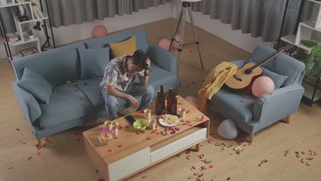 vista superior de un hombre asiático borracho sentado en un sofá y sosteniendo su cabeza con las manos después de tener una fiesta en casa