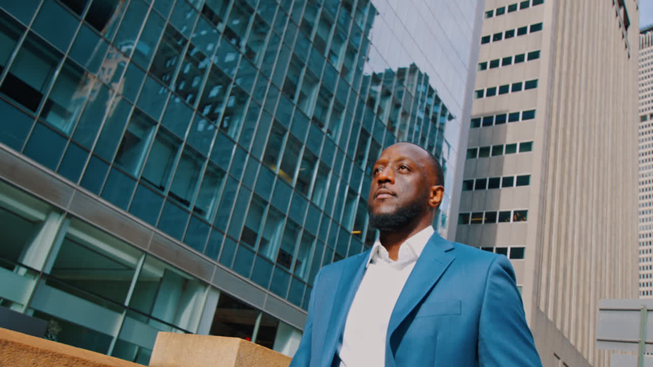 New York, USA, 1 April 2026: Businessman in blue suit beside glass office buildings. Low angle medium portrait of Black businessman in blue suit holding coat near glass towers in New York