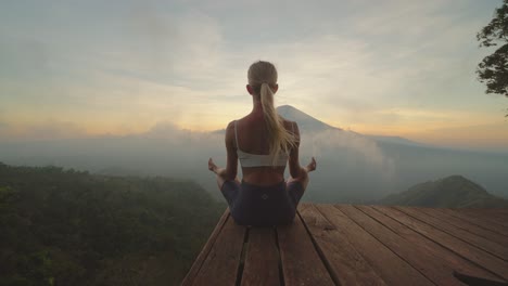 fit woman in sportswear meditating on platform with view of mount agung, sunset