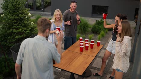 Vista-Trasera-De-Un-Chico-Rubio-Confiado-Con-Su-Compañía-Jugando-Beer-Pong-Y-Lanzando-Una-Pelota-Blanca-A-Un-Lado-Durante-Una-Fiesta-En-Una-Casa-De-Campo.