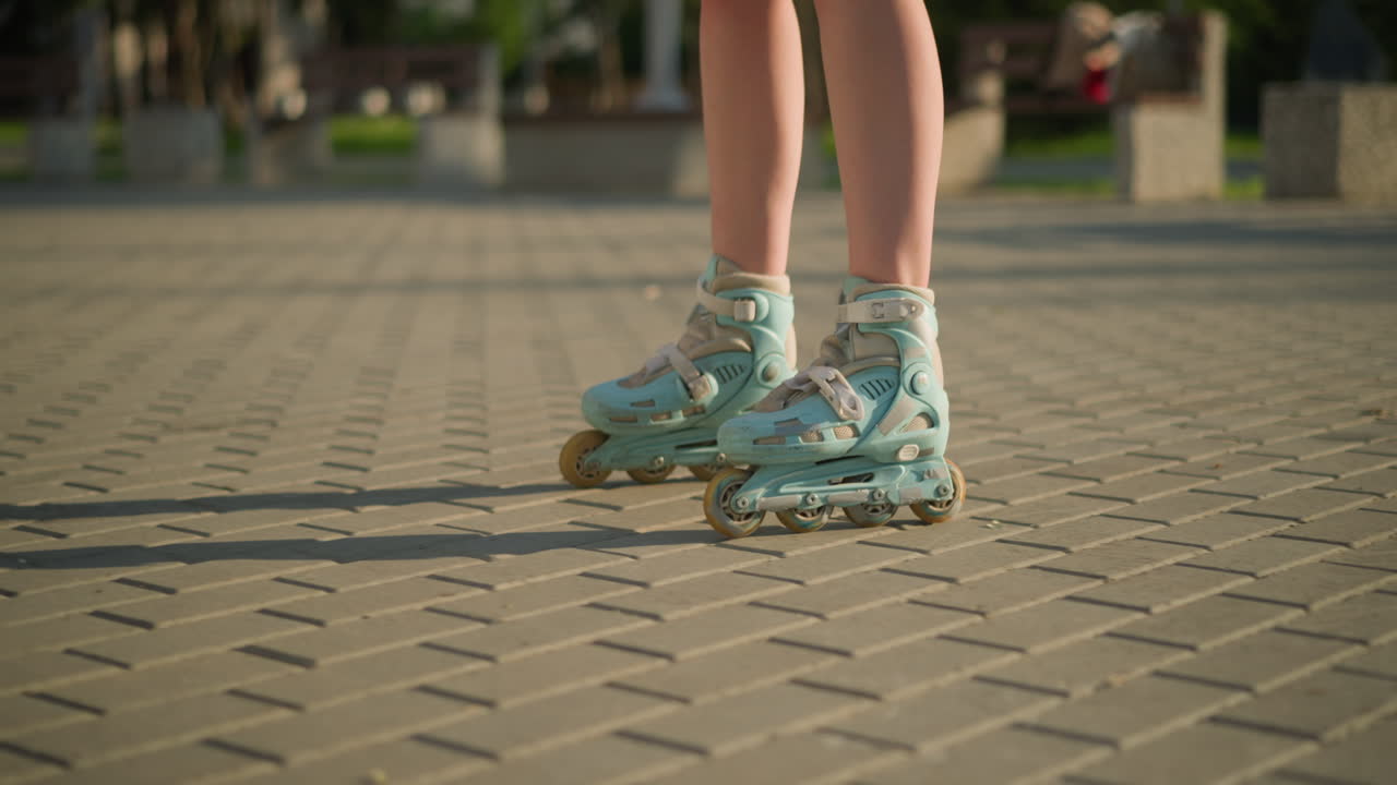 Leg view of person skating along interlocked path on rollerblades under warm sunlight casting shadows on ground, with blurred background featuring greenery and some structures