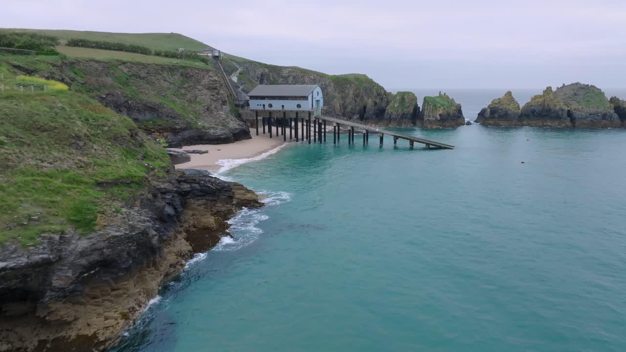 Jagged grassy cliffs next to clean blue sea with camera flight towards lifeboat station with slipway next to sea cliffs. Mother Ivey's Bay, Cornwall, UK.