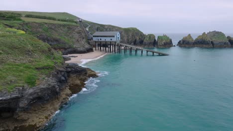 jagged grassy cliffs next to clean blue sea with camera flight towards lifeboat station with slipway next to sea cliffs. mother ivey's bay, cornwall, uk.