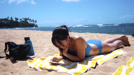 wide slow-motion shot of a young asian woman scrolling on mobile device and sunbathing at idyllic beach