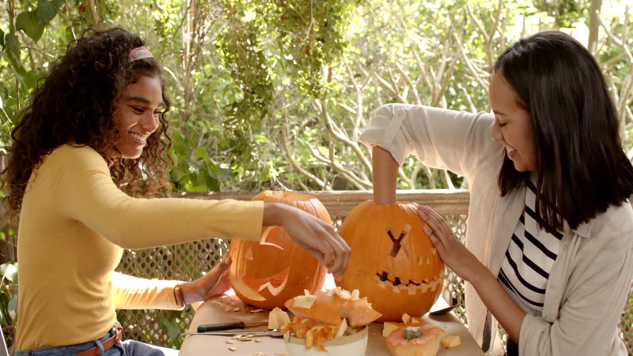 Halloween time, two multiracial female friends carving pumpkins on a porch, at home