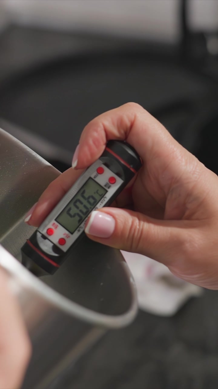 Primo piano di una mano che tiene un termometro digitale per misurare la temperatura di un liquido in un contenitore. Le unghie della persona sono ben curate con smalto rosa chiaro. L'illuminazione soffusa evidenzia i dettagli di entrambi.