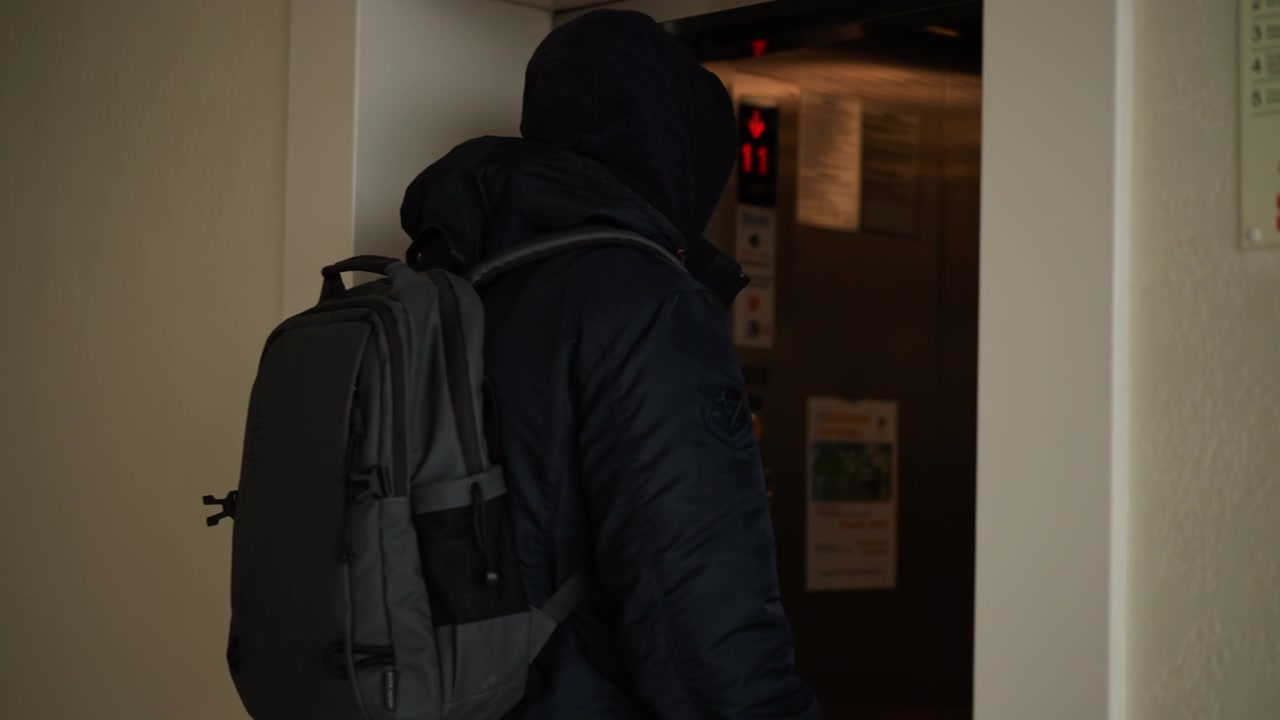 Primo piano di un uomo in abiti scuri che entra in un ascensore con le porte chiuse, portando uno zaino grigio. La scena mostra un interno metallico e un display digitale che indica il piano 11 mentre l'uomo entra.