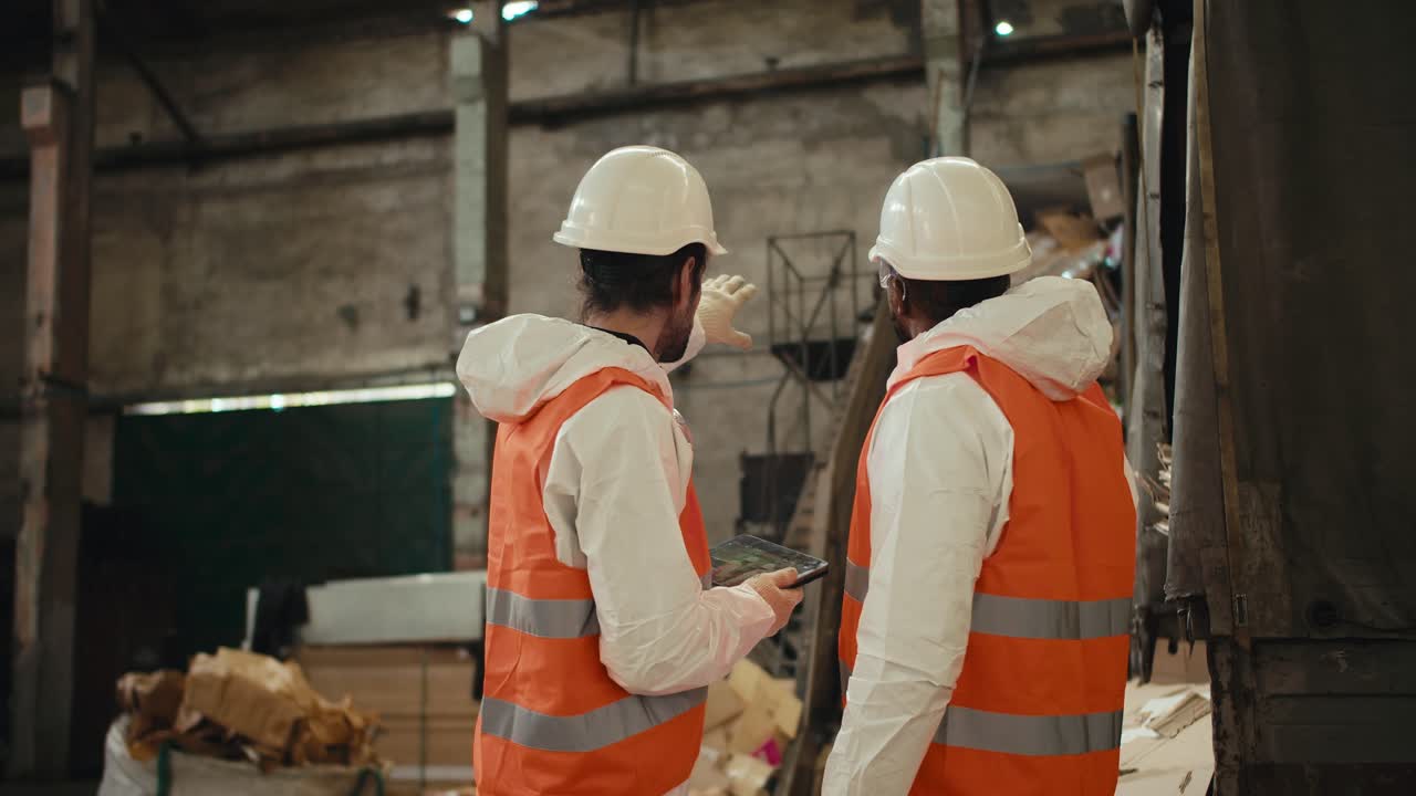 Rear view of a duo of men in special protective clothing and orange vests talking and discussing How to better design plans at their waste recycling plant while recycling waste paper on a conveyor belt